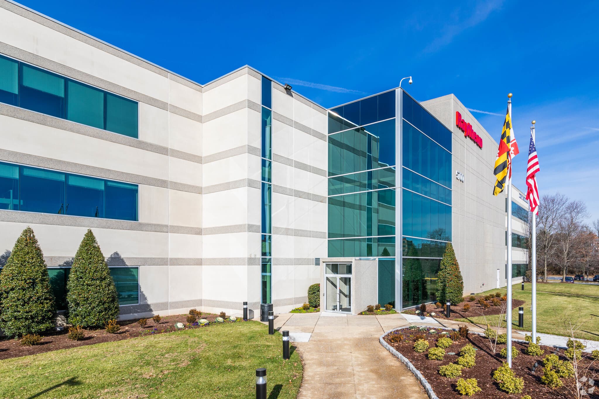 Front entrance to 5700 Rivertech Court, in College Park, MD. Maryland and American flags sit in front of a large glass entrance to the building