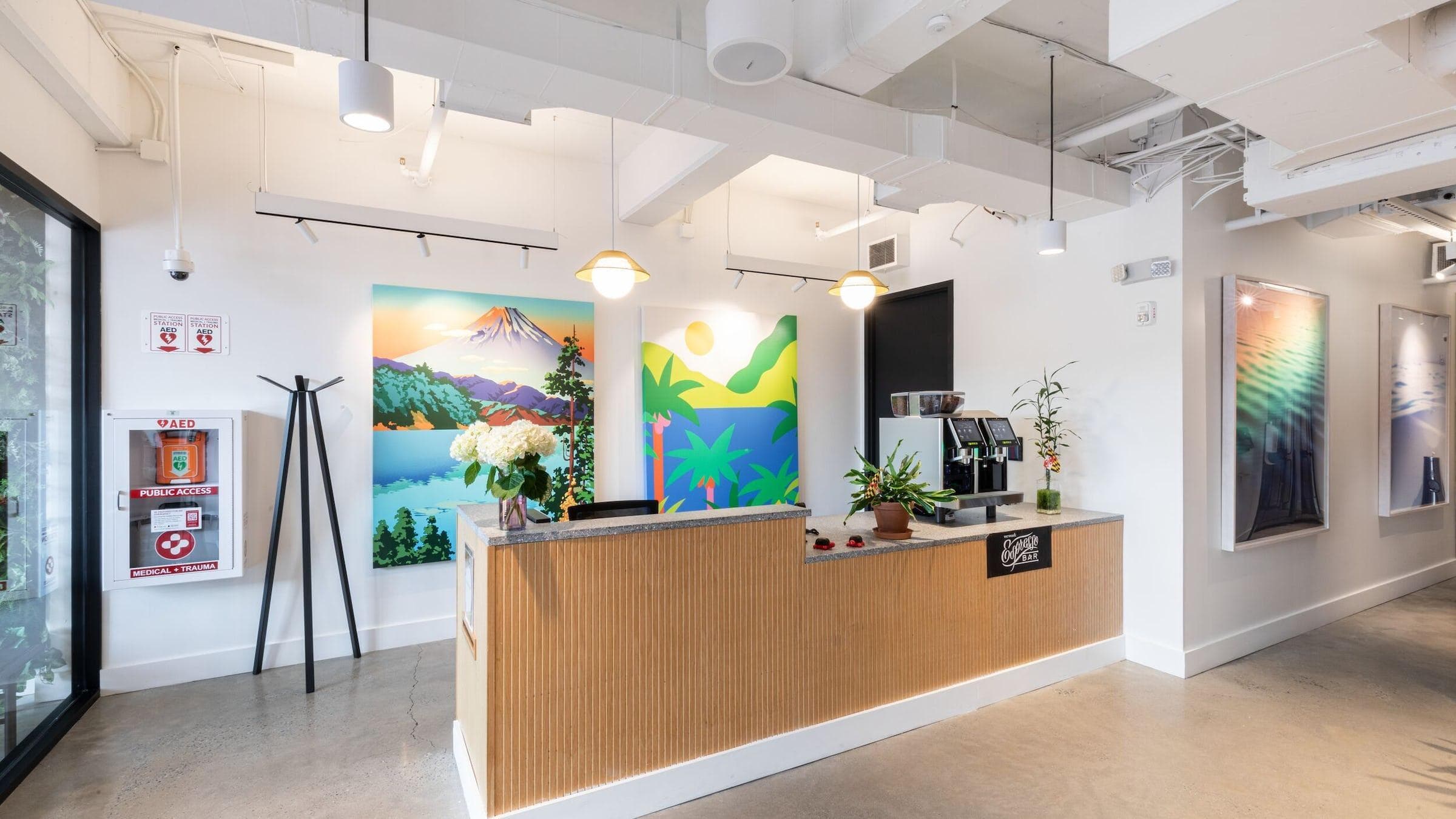 Entrance hall to WeWork in College Park. A welcome desk flanked by a coffee machine station. Green office plants dot the space with Abstracted landscape painting along the walls.