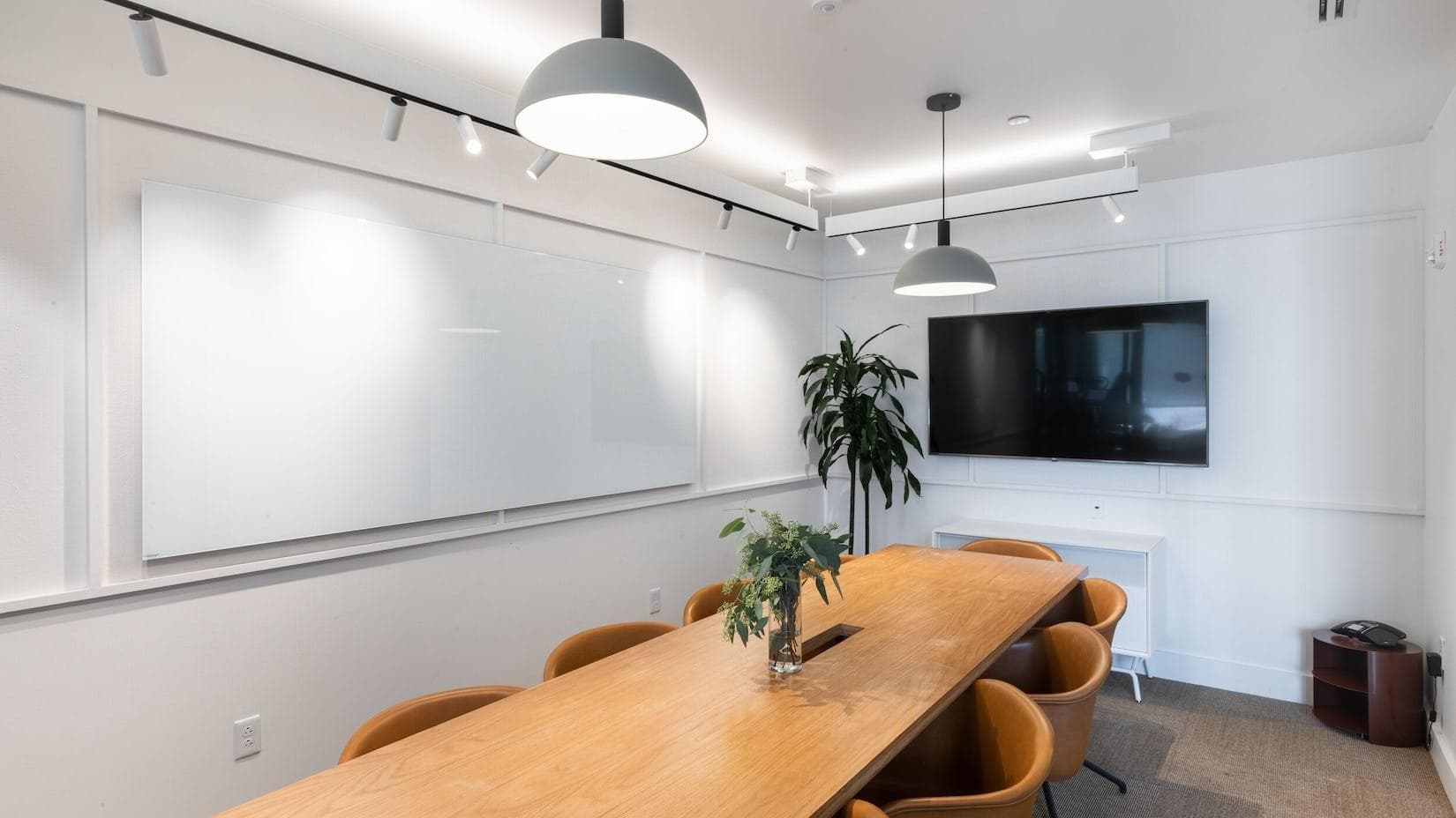 A Conference room in the College Park WeWork space. Clean dry erase boards, a TV screen and some office plants decorate the space along with the table and brown leather chairs.
