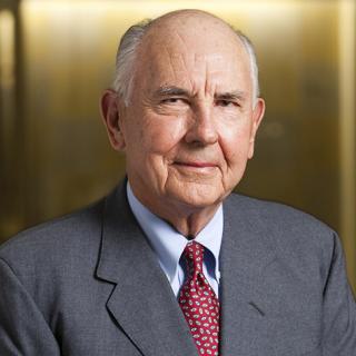 Headshot of A. James Clark, smiling man with white hair, gray suit, red tie, light blue dress shirt, and gold background.