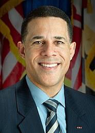 Full color headshot of Anthony Brown wearing a navy blazer and blue dress shirt against a background of flags.