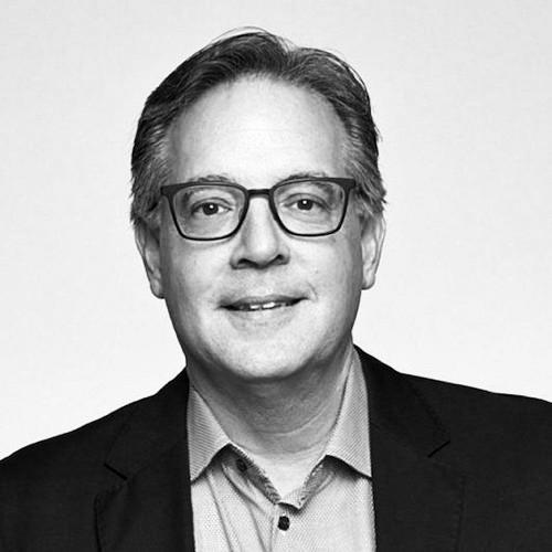 Black and white headshot of Dan Millman smiling and wearing a dark blazer and dress shirt