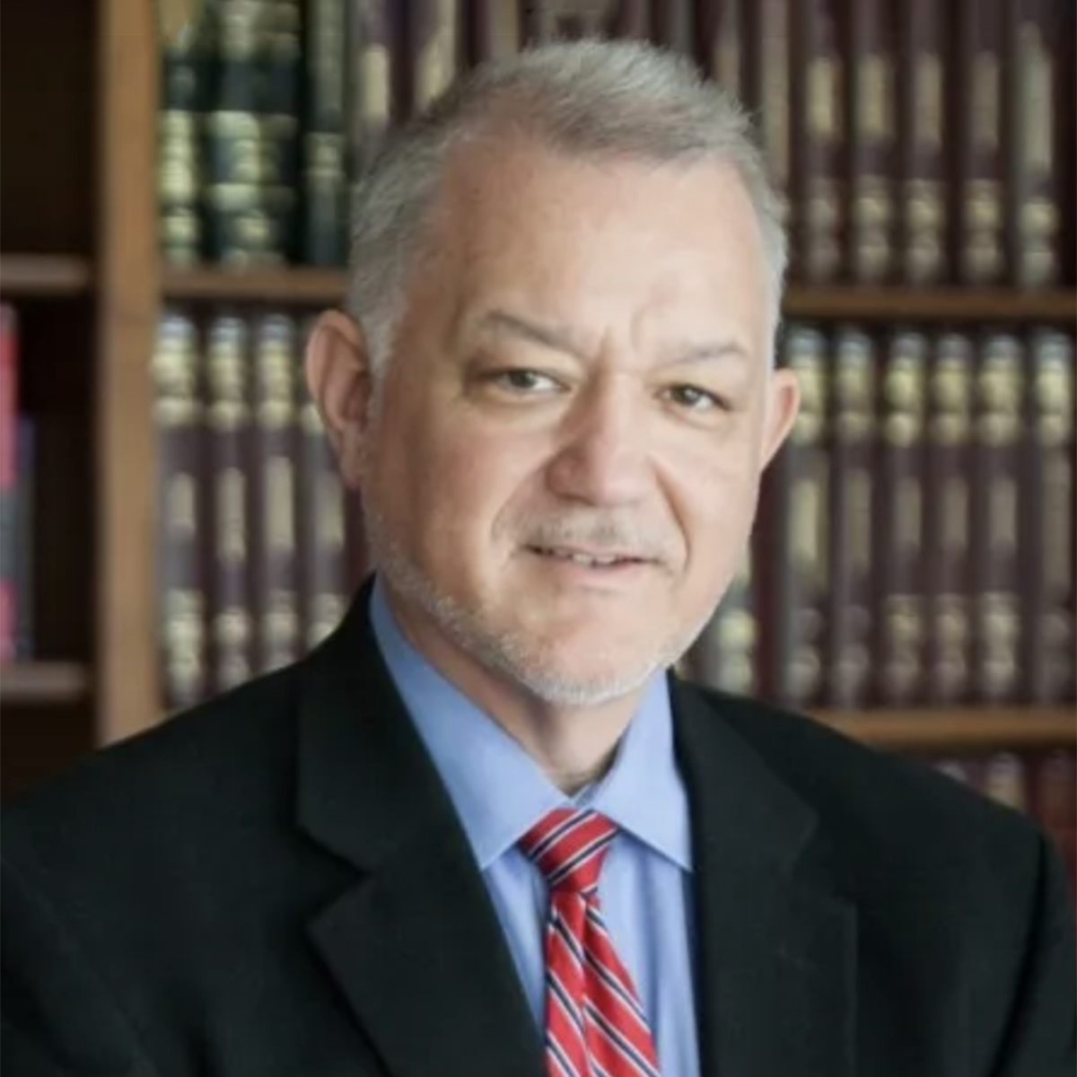 Full color of David Brinkley smiling wearing a black blazer, blue dress shirt, and red striped tie with a bookcase in the background