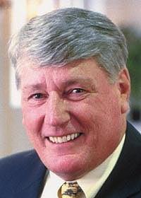 Mike Busch smiles wearing a black blazer, lapel pin, yellow shirt, and yellow tie.