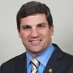 Full color headshot of Douglas J.J. Peters smiling against a gray colored background with a black blazer, lapel pin, blue dress shirt, and striped tie.
