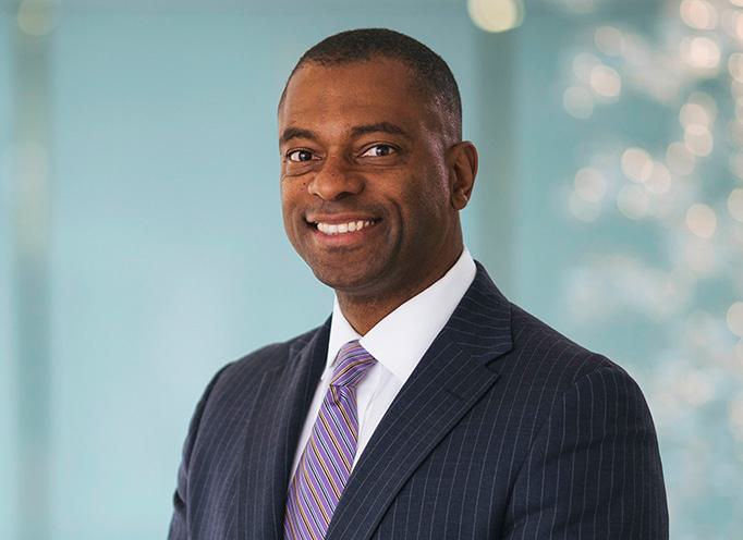 Full color headshot of Craig Thompson smiling wearing a navy pinstripe suit, white dress shirt, and striped tie against a light turquoise background.